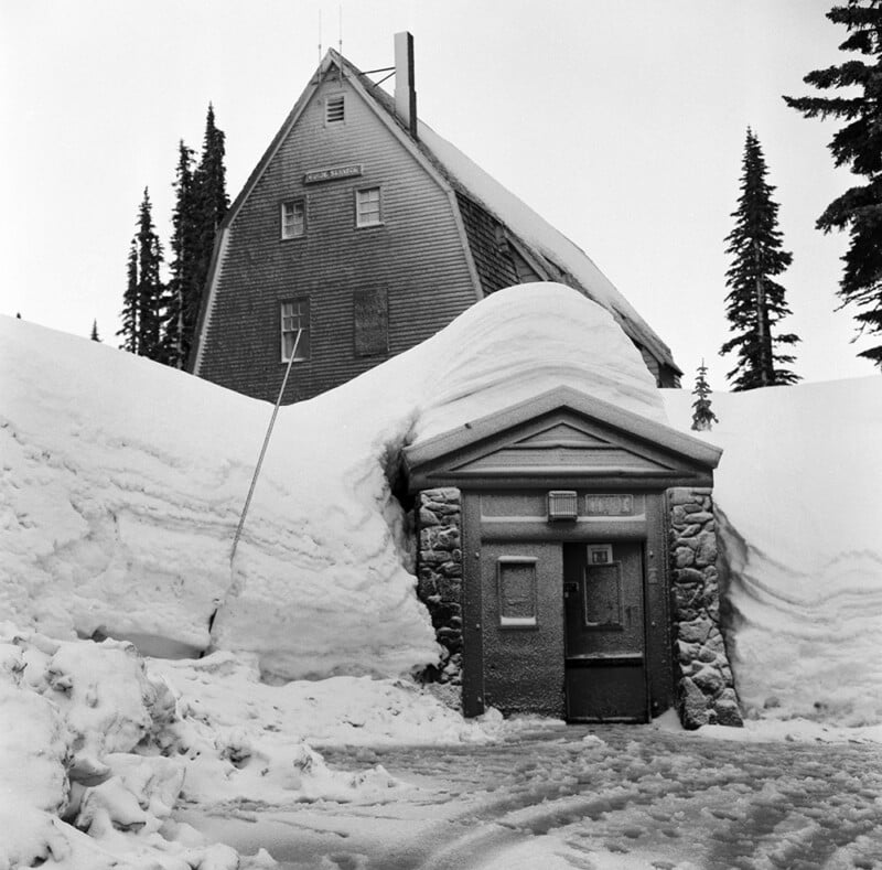A large, wooden, multi-story building with a steep roof stands behind a stone-framed entrance buried in deep snow.