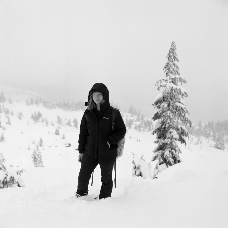 A person wearing a hooded black jacket and dark pants stands in a snowy, mountainous landscape next to a snow-covered tree.