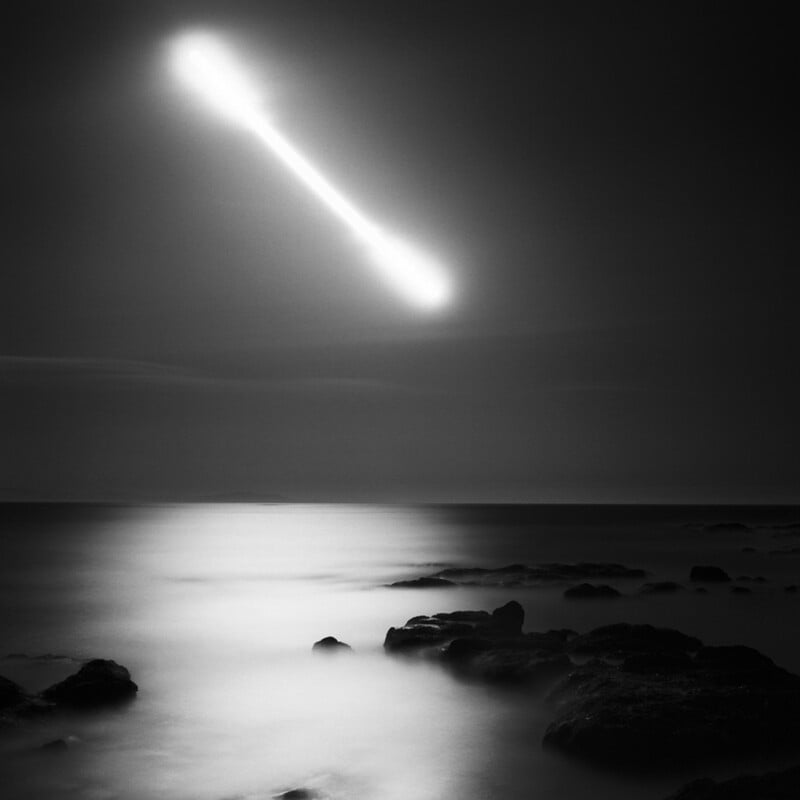 A long exposure, black-and-white photograph shows a bright, streaking light in the night sky above a calm, rocky coastline.