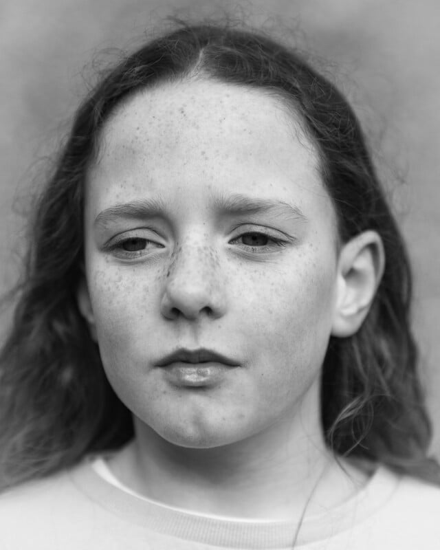 A black and white close-up portrait of a young person with freckles, long wavy hair, and a neutral expression.