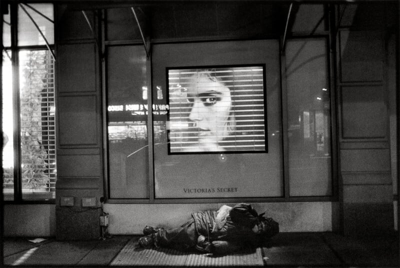 A person sleeps on a sidewalk in front of a storefront window displaying a large, backlit portrait of a woman's face behind security shutters.