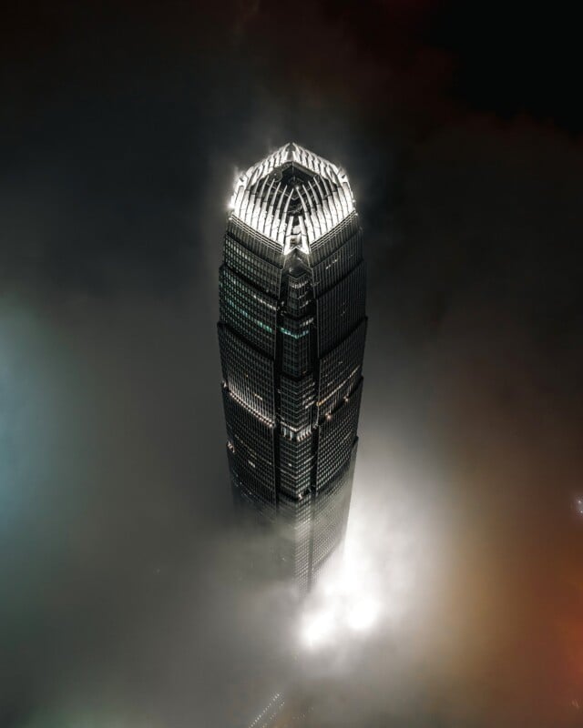 A skyscraper rises above a thick layer of low-hanging clouds at night, with its illuminated crown glowing against the dark sky.