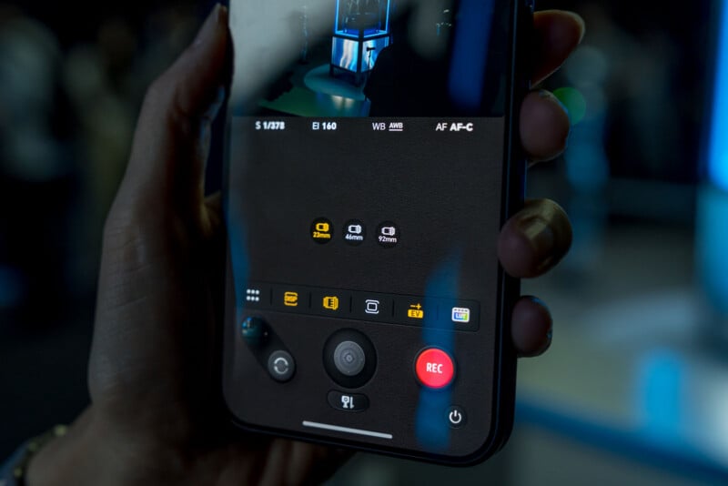 A close-up view of a hand holding a smartphone displaying a professional camera interface with manual settings, lens options, and a red record button.