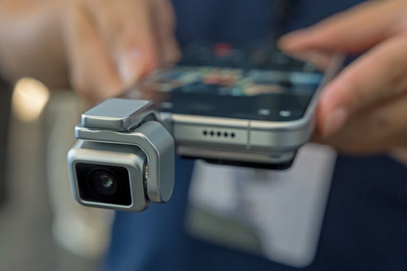 A close-up view of a person holding a smartphone with a small, silver, gimbal-mounted camera attached to the side.