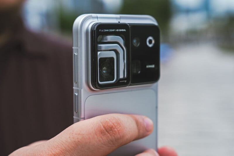 A close-up of a hand holding a silver smartphone, focusing on the large, complex camera module with text reading "F1.6 23MM 200MP / 4D GIMBAL".