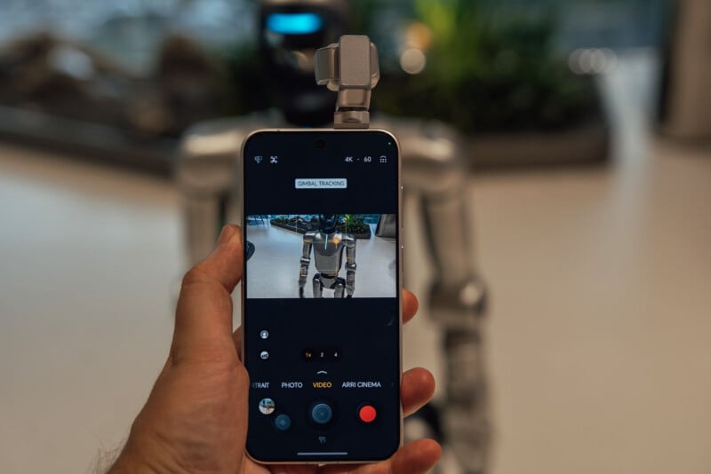 A person holds a smartphone displaying a camera interface with "GIMBAL TRACKING" enabled, pointed at a metallic humanoid robot.