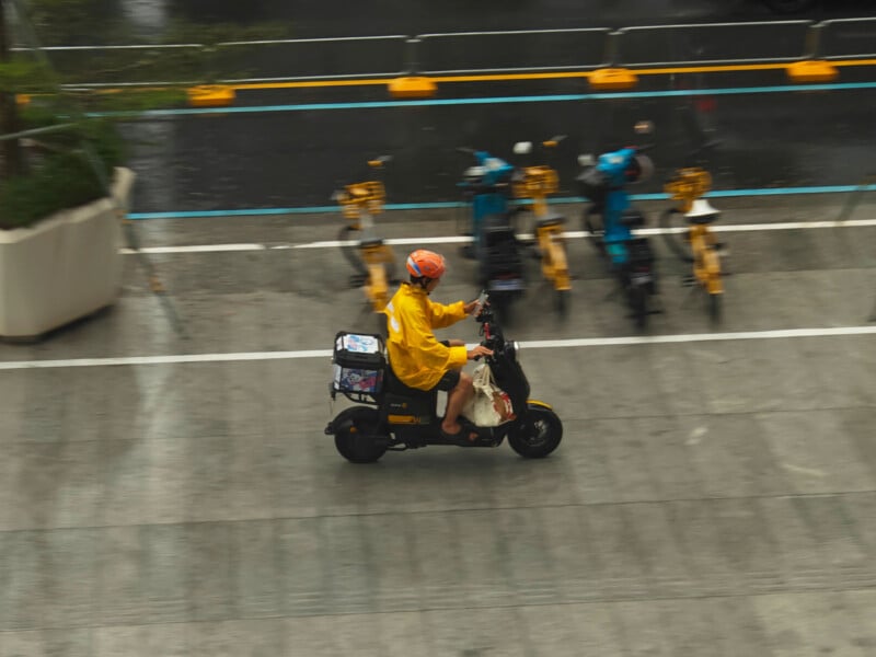 A delivery driver in a yellow raincoat and orange helmet rides a black electric scooter past a row of parked scooters on a wet street.