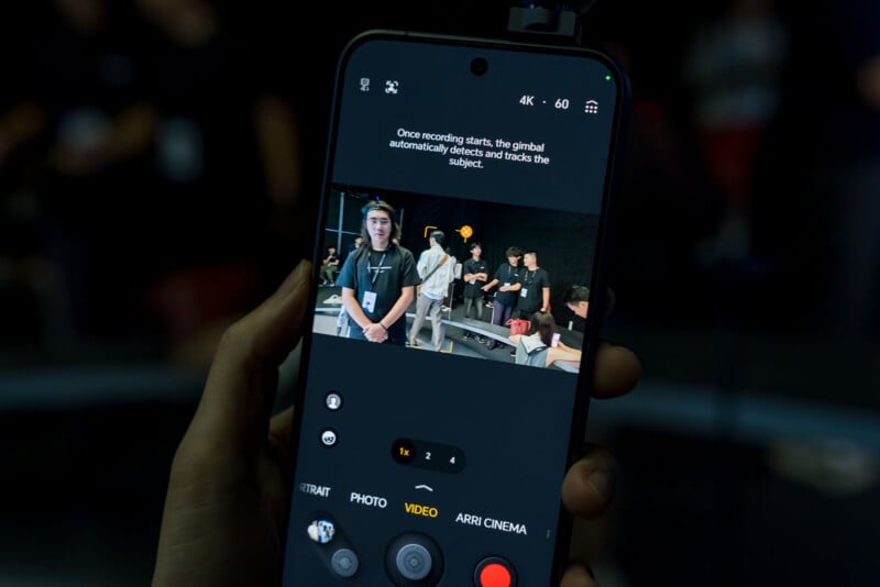 A hand holds a smartphone displaying a camera interface with a gimbal tracking feature active, showing a group of people in a studio setting.