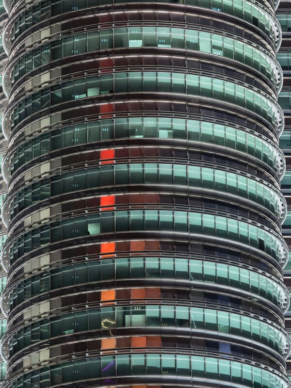 A close-up view of a cylindrical skyscraper facade featuring horizontal bands of glass windows, with a vertical column of orange-lit offices visible in the center.