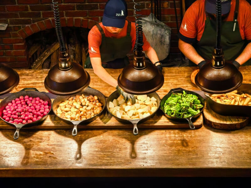 Two chefs in aprons and hats prepare food in cast-iron pans under heat lamps in a kitchen with a brick fireplace.