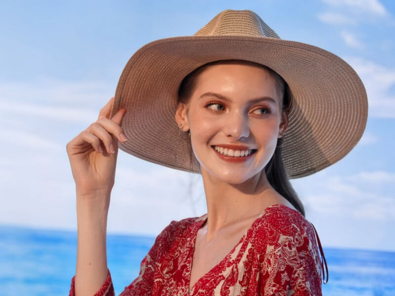 A smiling woman wearing a wide-brimmed straw hat and a red patterned dress looks to the side against a blue sky and sea background.