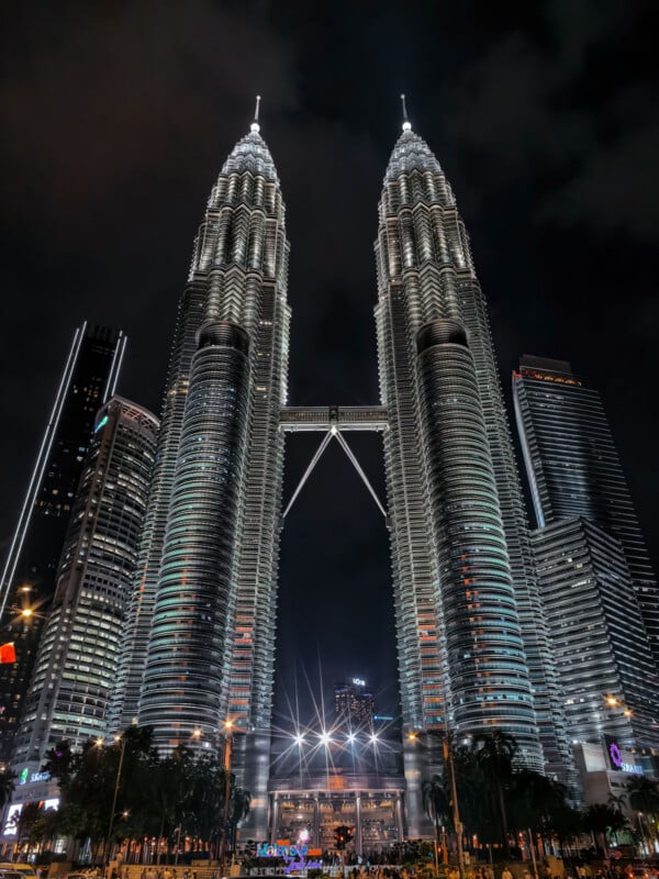 The Petronas Twin Towers are illuminated at night, connected by a skybridge, with surrounding skyscrapers and a park visible at the base.