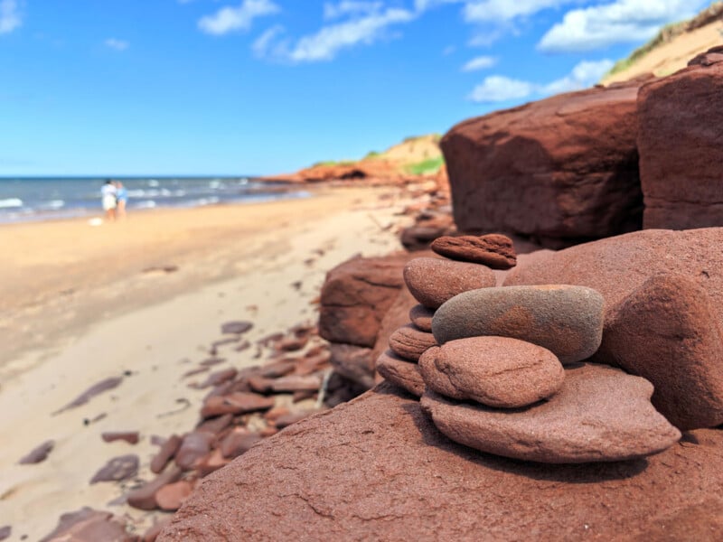 A stack of small, smooth red stones rests on a larger rock formation overlooking a sandy beach and the ocean under a blue sky.