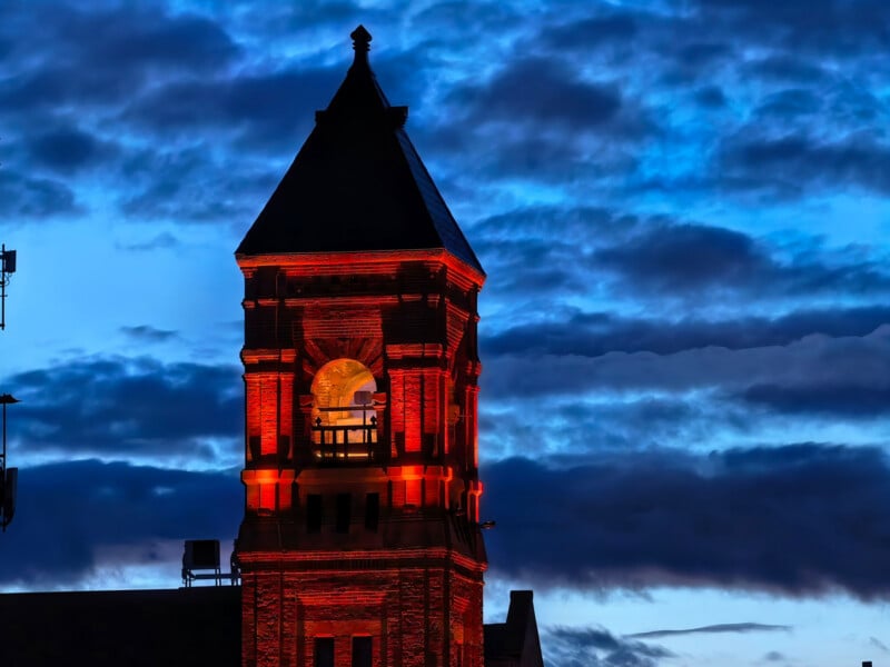 A brick clock tower illuminated in red light stands against a dark, cloudy blue evening sky.