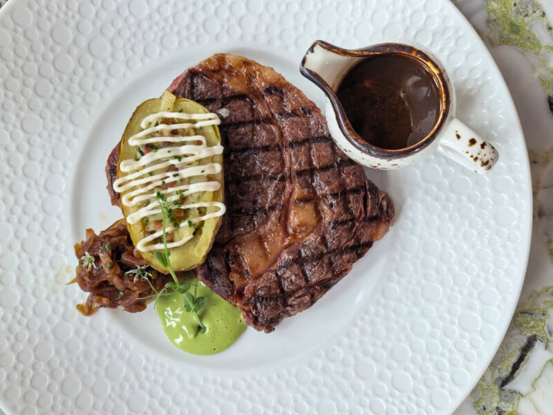 A grilled steak served with a stuffed potato, caramelized onions, a dollop of green sauce, and a small pitcher of gravy on a white textured plate.