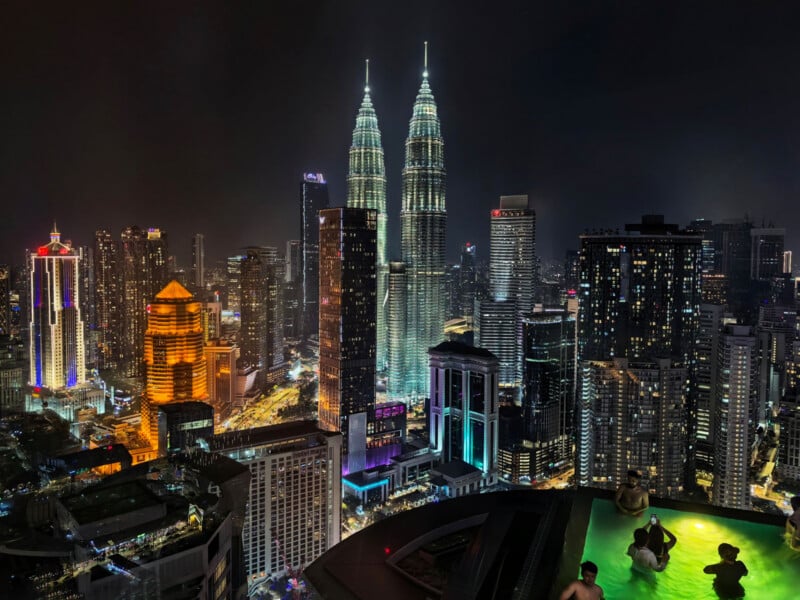 A nighttime cityscape features the illuminated Petronas Towers rising above a dense skyline, with people swimming in a rooftop infinity pool in the foreground.
