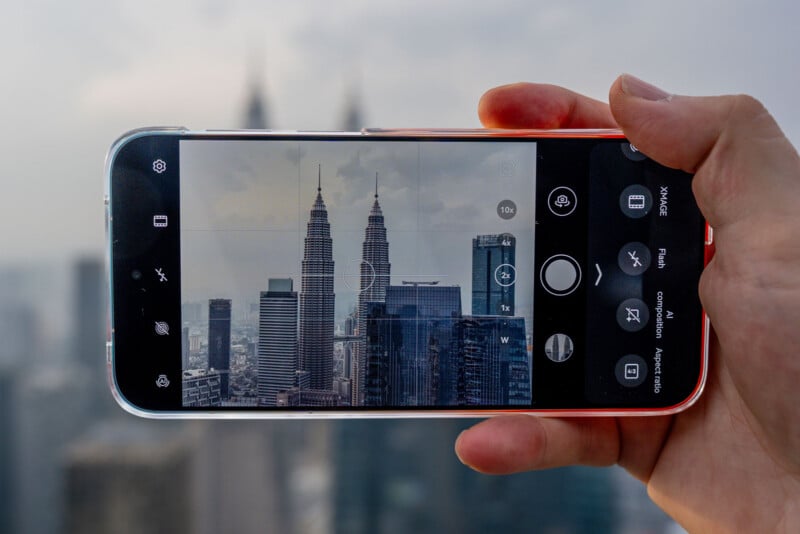 A hand holds a smartphone displaying a camera interface, showing a view of two tall skyscrapers in a city skyline.