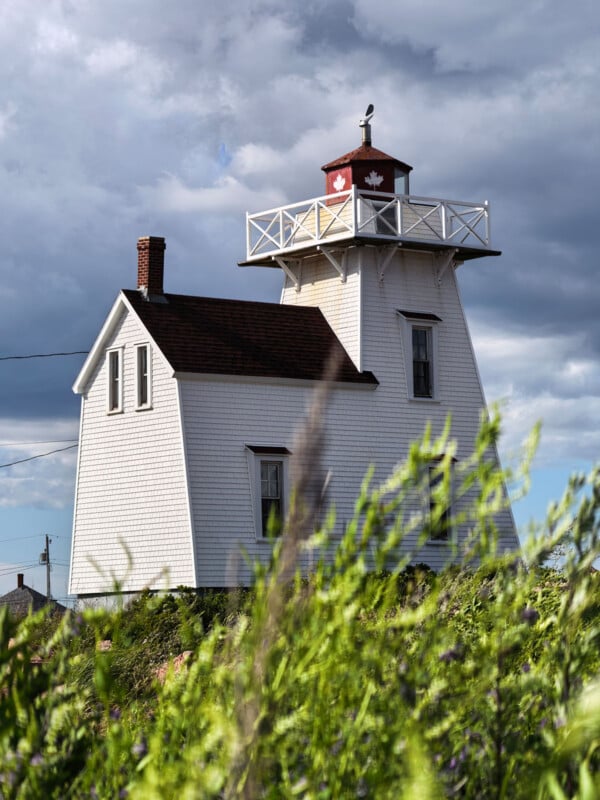 A white wooden lighthouse with a red roof and Canadian maple leaf accents stands behind a foreground of blurred green grass.