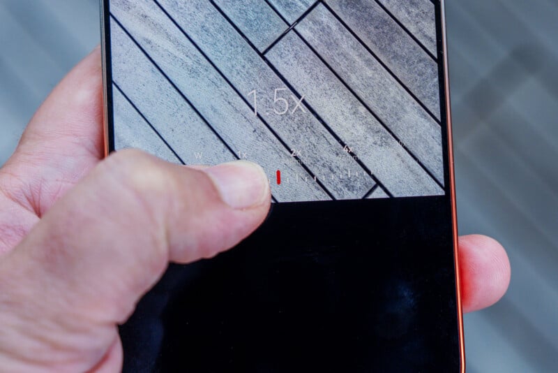 A hand holds a smartphone displaying a camera zoom interface set to 1.5x magnification over a background of diagonal wooden planks.