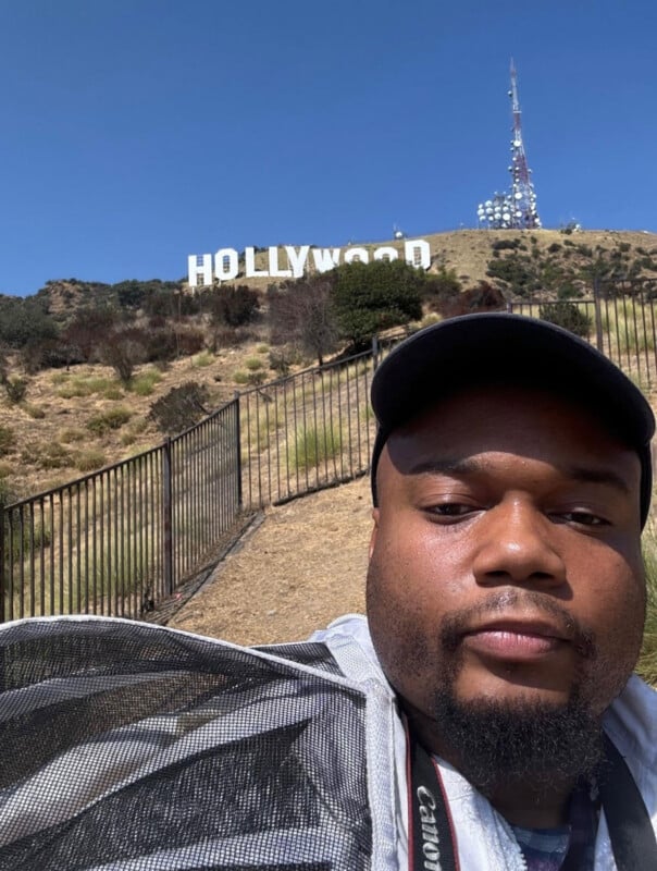 A man wearing a black baseball cap and a white mesh jacket takes a selfie in front of the Hollywood sign on a sunny day.