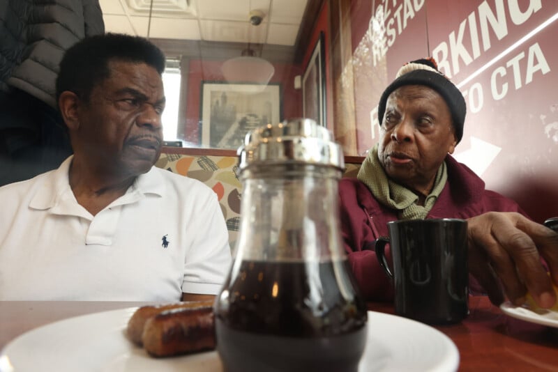 Two older Black men sit at a restaurant table, looking at each other while a syrup dispenser sits in the foreground.