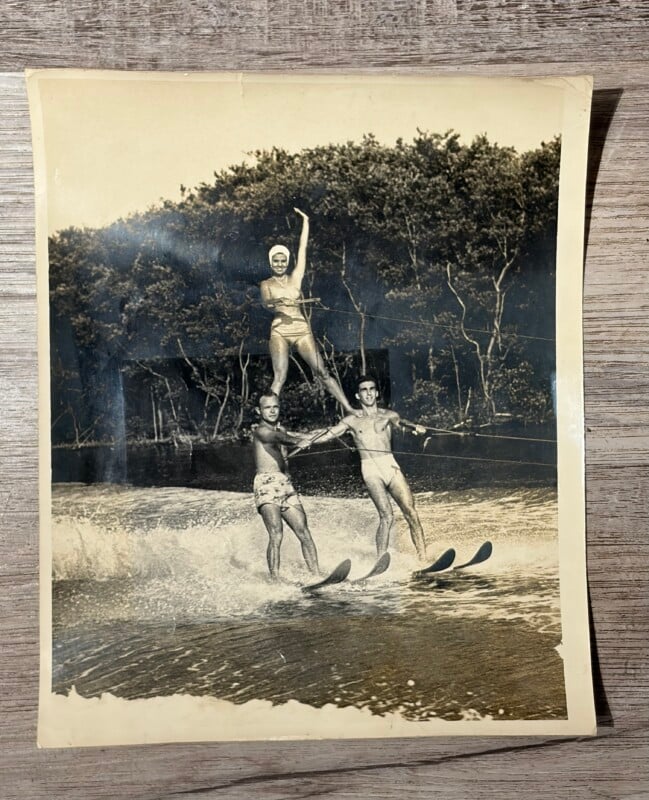 A woman in a swim cap and swimsuit stands on the shoulders of two men as they all water ski together on a lake.