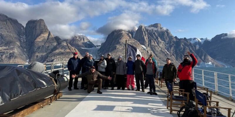 A group of people wearing eclipse glasses stand on the deck of a boat in front of a large, mountainous, snow-capped landscape.