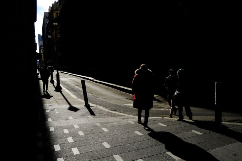 Pedestrians walk along a city sidewalk in high-contrast sunlight, casting long shadows against a dark building facade.