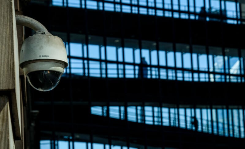 A Bosch security camera is mounted on a wall in front of a modern building with a glass facade and visible interior walkways.