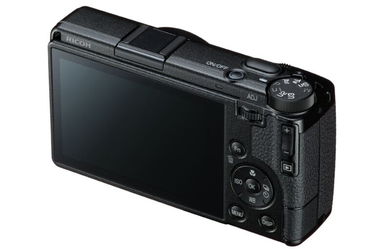 A black Ricoh digital camera viewed from the rear, showing the LCD screen, mode dial, and various control buttons.