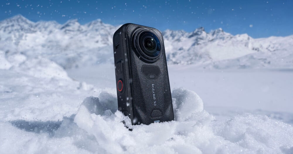 An Insta360 X4 camera stands upright in a snowbank against a backdrop of snow-covered mountains under a clear blue sky.