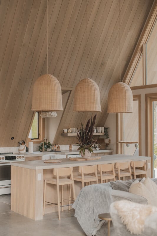 A kitchen island with five light-wood stools sits beneath three large, woven pendant lights in an A-frame room with wood-paneled walls.