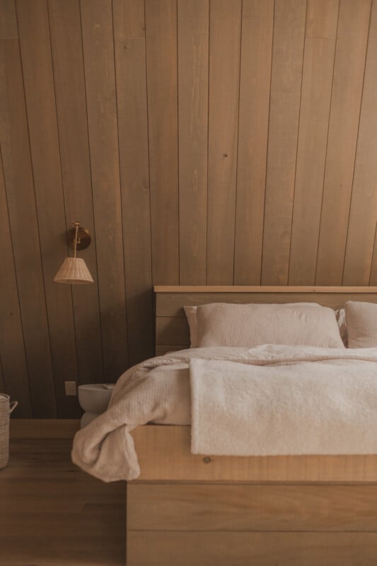 A bedroom features a wooden bed frame with beige bedding against a vertical wood-paneled wall, illuminated by a wall-mounted sconce.