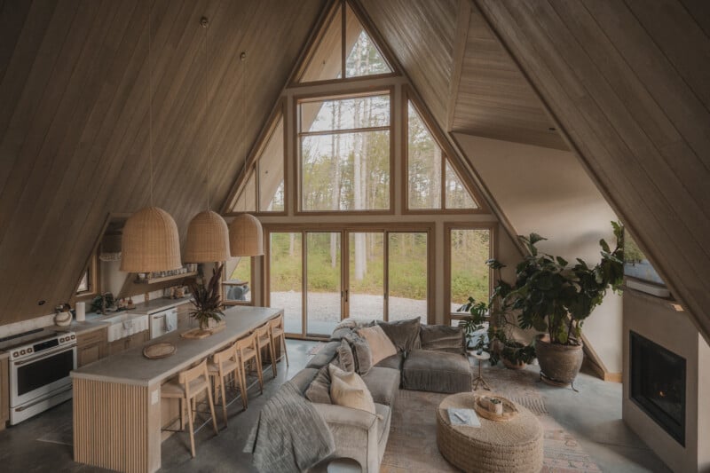 An A-frame cabin interior features an open-concept kitchen with a breakfast bar, a living area with a sectional sofa, and floor-to-ceiling windows overlooking a forest.