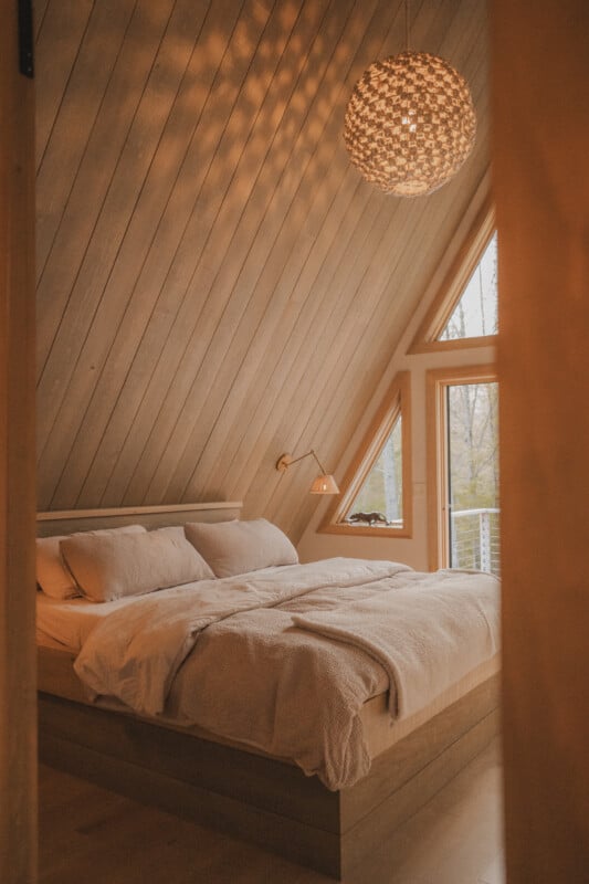 A cozy bedroom in an A-frame cabin features a platform bed with neutral bedding, a textured pendant light, and a window overlooking a forest.