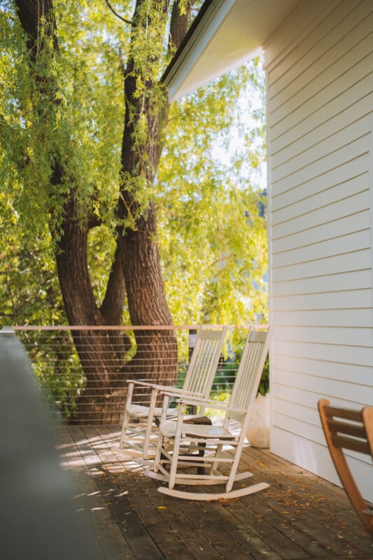 Two white wooden rocking chairs sit on a wooden deck next to a white-sided house, with a large tree in the background.