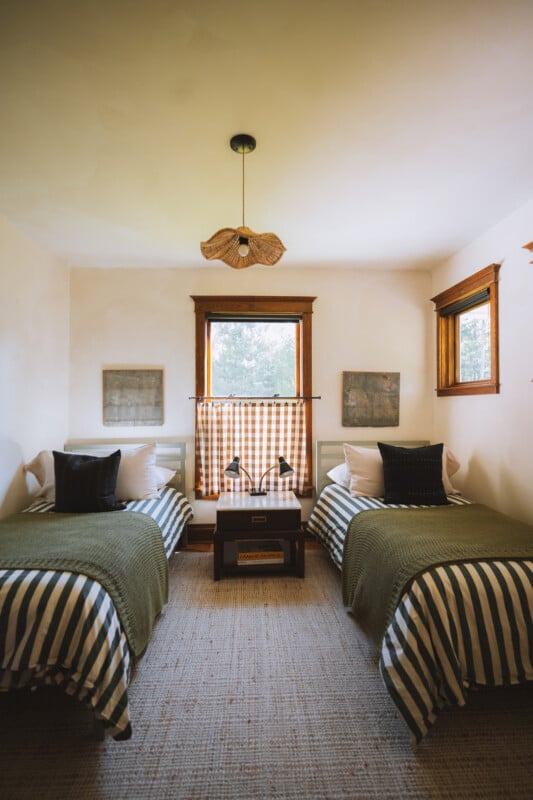 A bedroom features two twin beds with striped bedding and green throws, separated by a small nightstand with a double lamp under a window with a gingham curtain.