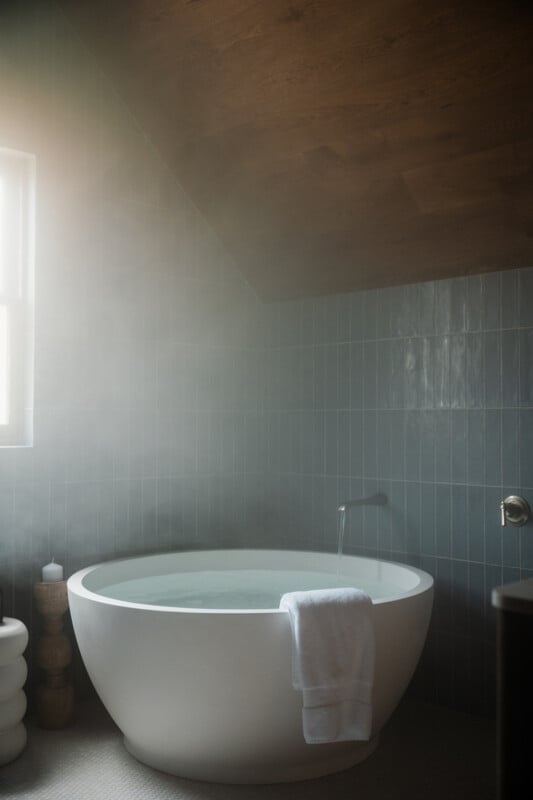A white circular bathtub filled with water sits in a tiled bathroom with a wooden ceiling and steam rising from the water.