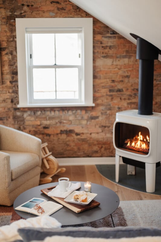 A cozy room features a white wood-burning stove, a beige armchair, and a round coffee table with a book, a mug, and a pastry on a wooden board.