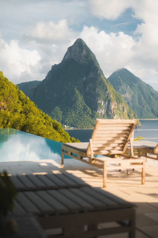 An infinity pool overlooks two large, lush green mountains rising from the sea, with wooden lounge chairs on the deck.