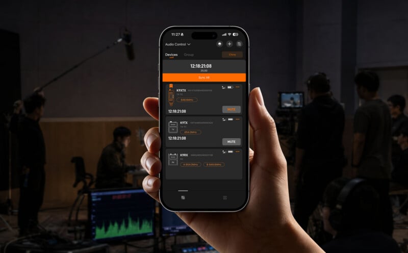 A hand holds a smartphone displaying an audio control app with a list of connected wireless transmitter and receiver devices.