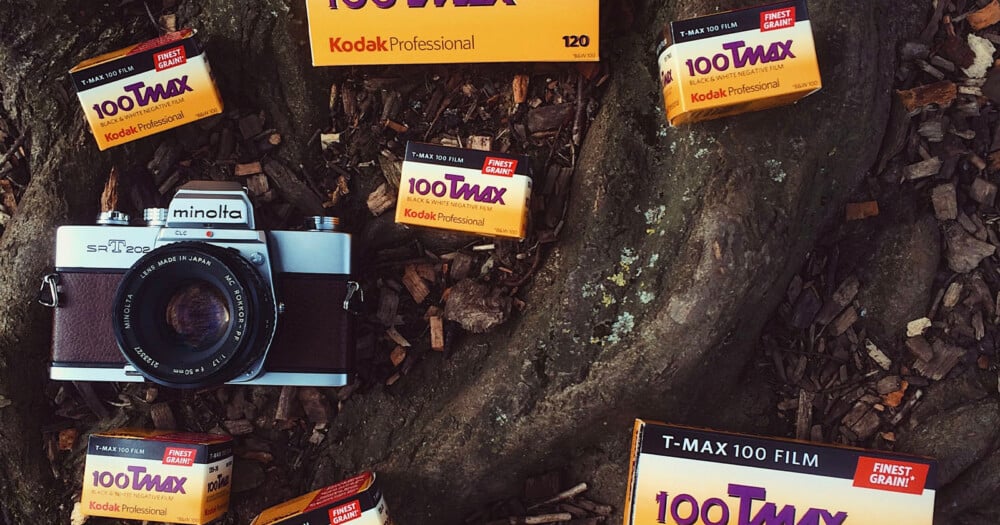 A Minolta SRT 202 camera sits on a tree root surrounded by several boxes of Kodak Professional T-MAX 100 film.