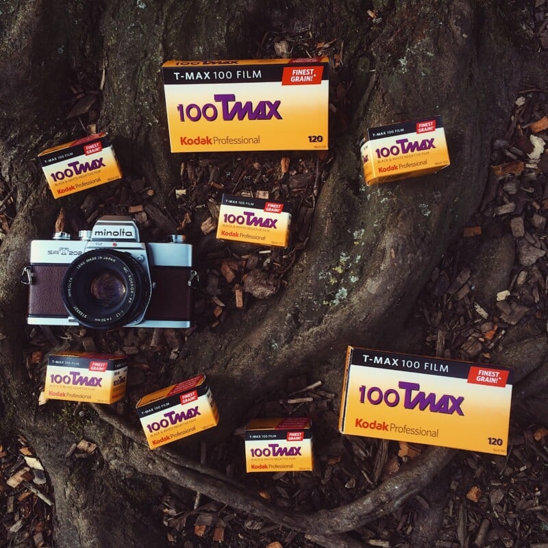 A Minolta SRT 202 camera surrounded by several boxes of Kodak Professional T-MAX 100 film on a tree root background.