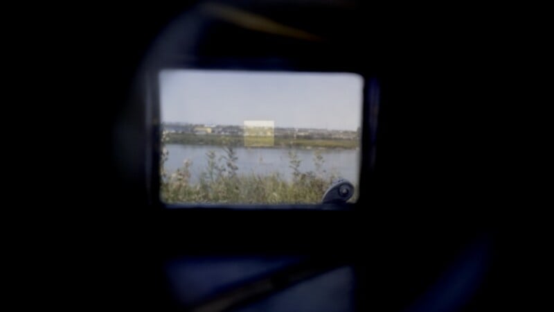 A view through a camera viewfinder showing a riverbank with tall grass, a body of water, and distant buildings under a clear sky.