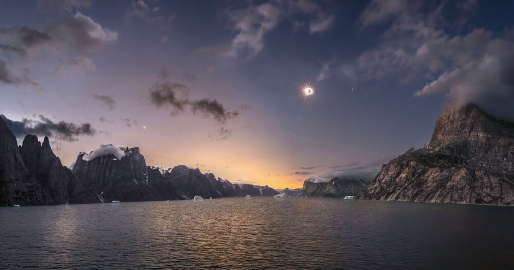 A solar eclipse shines above a calm body of water surrounded by jagged, rocky mountains under a twilight sky.