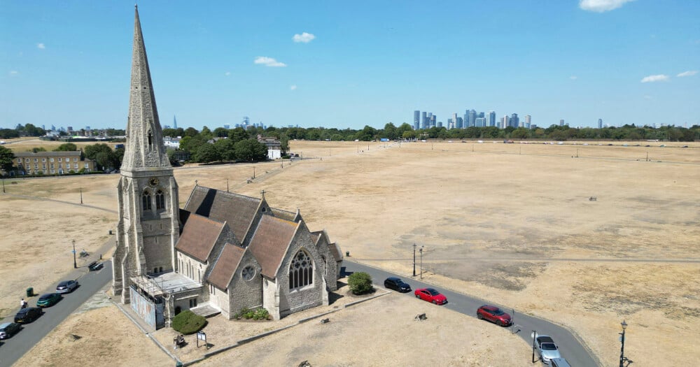 An aerial view of a stone church with a tall spire situated next to a large, dry grassy field, with a city skyline visible in the distance.