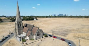 An aerial view of a stone church with a tall spire situated next to a large, dry grassy field, with a city skyline visible in the distance.