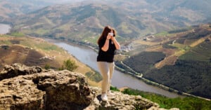 A woman stands on a rocky cliff edge, holding a camera to her face to photograph the river valley below.