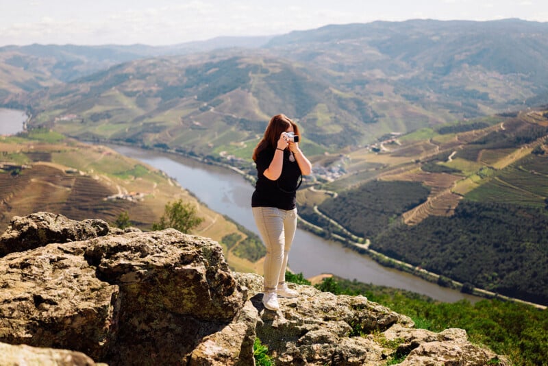 A person with long brown hair stands on a rocky cliff edge, holding a camera up to their face to take a picture of the winding river and terraced hills below.
