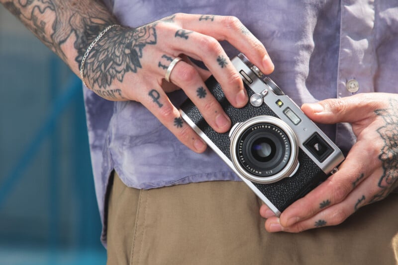 A person with tattooed hands holds a silver and black Fujifilm camera against a light purple shirt and tan pants.